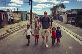 In the background blue sky. Left and right of a small street are colourful huts. In the middle of the street a young man with sunglasses is walking. At each of his hands, he has 2  little children at his hands.